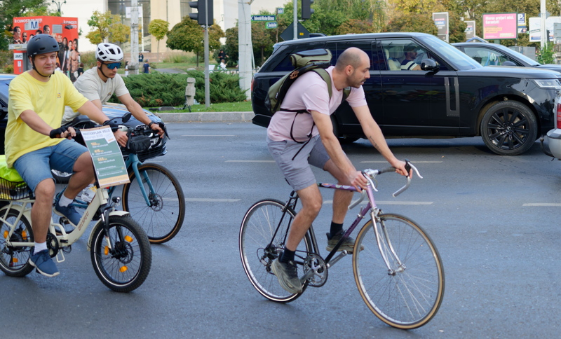 5 - Three people riding bicycles on a paved urban road near traffic and a black SUV